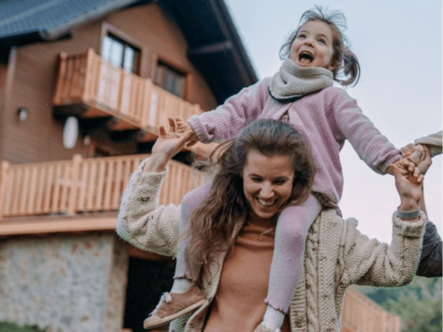 A woman with a child on her shoulders smiling. In the background is a house with solar panels